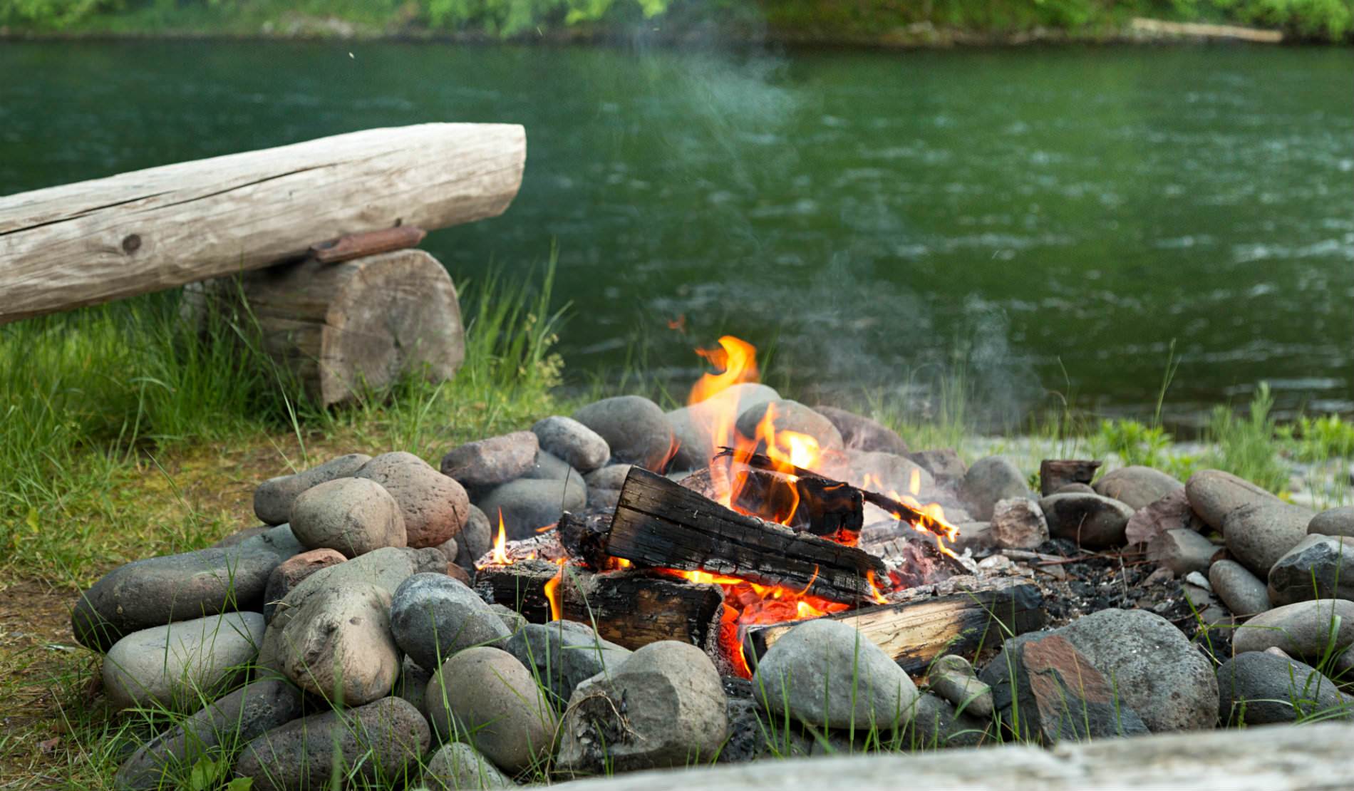 An orange fire glowing in a smooth rock lined firepit near a wood bench and a river.