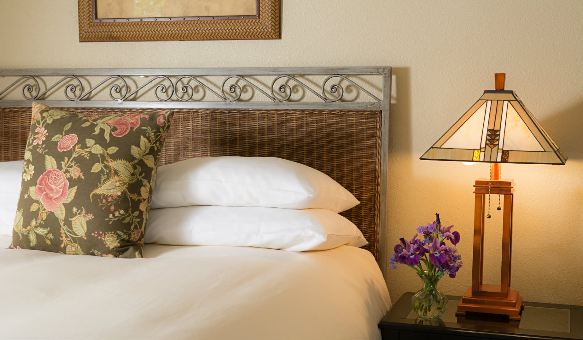Soft white bedding and pillows on bed next to sidetable with craftsman-style lamp and purple flowers.