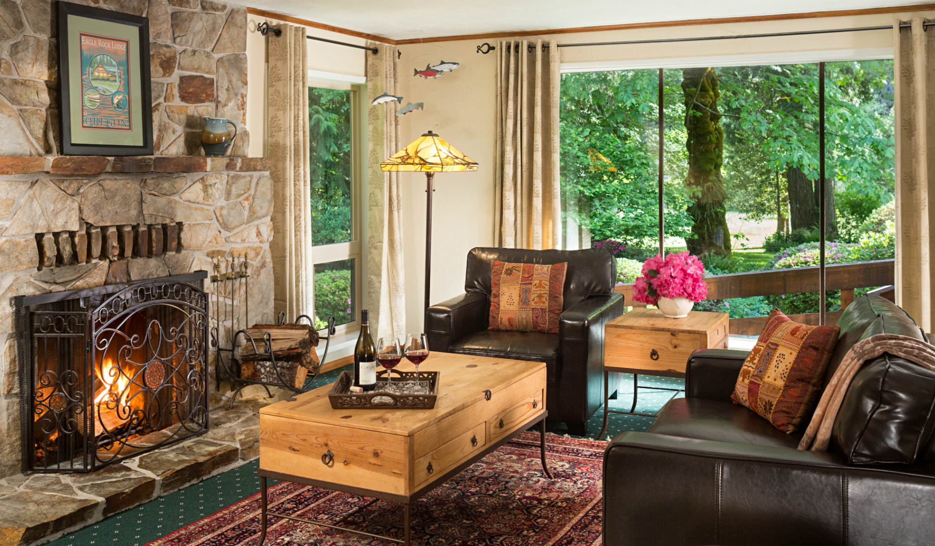 Large stone fireplace facing two leather club chairs with large windows viewing greenery outside.