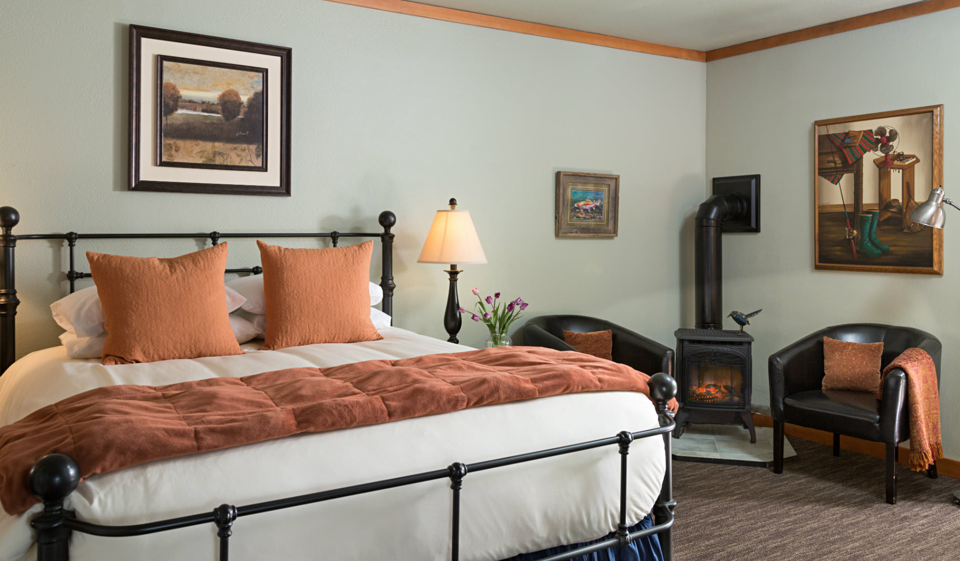 Large iron bedstand with soft rust and white bedding, two chairs beside wood burning stove.