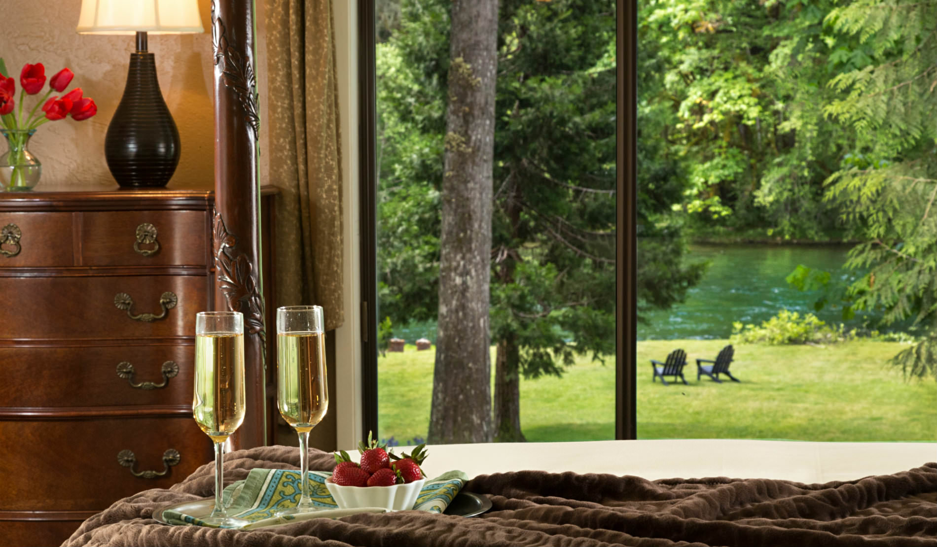 View from bed with two glasses of champagne and strawberries to brown dresser and grassy area and river, two chairs.