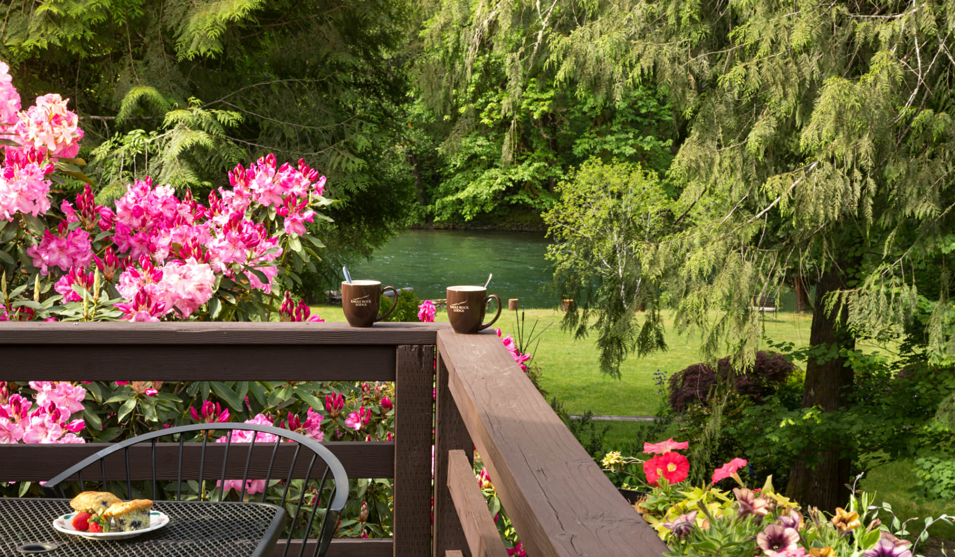 Corner of wooden deck with coffee mugs on ledge, pink flowers and green trees in background.