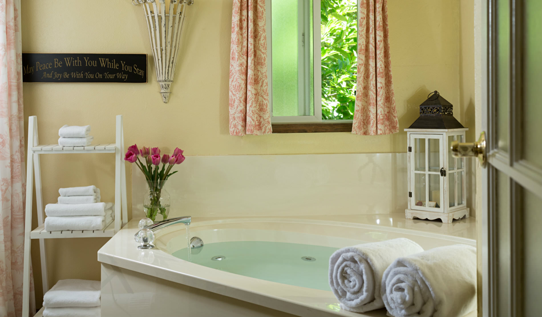 Whirlpool bath with white towels, pink flowers and window open to green tree.