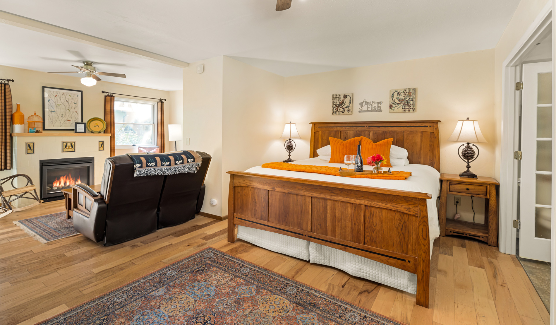 Wood Floored room with soft seating in front of fireplace, large wooden bed and soft bedding, beige walls.