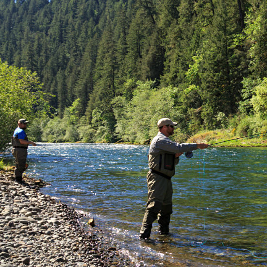 Two men fly fishing on the stony bank of the river with woods on the far bank