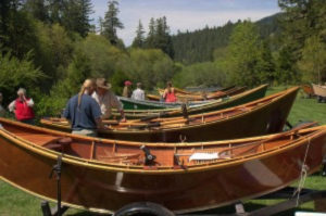 A group of people mill about a collection on wooden boats on trailers with lush green trees behind them. 